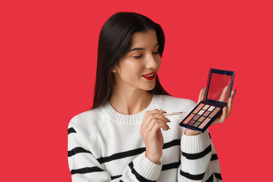 Beautiful young woman with brush and palette of eyeshadows on red background