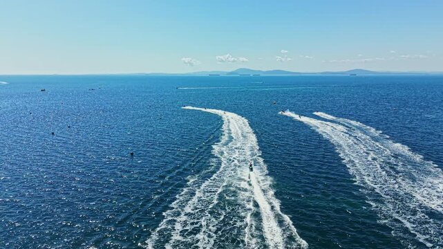 Riders race their jet skis across the water, creating trails behind them. The sky is blue with a few clouds as boats pass in the distance. Waves splash and spray around them.