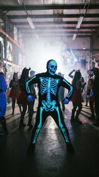 Halloween A person in a glowing skeleton costume strikes a dramatic pose in the center of a hazy warehouse party, with costumed figures dancing in the background under dramatic lighting.