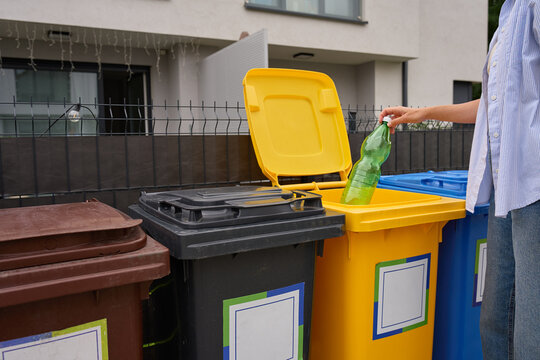 Person placing plastic bottle into yellow recycling bin outside residential home