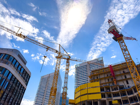 Cranes on a site, blue sky. Cranes stand tall at a construction site in a city during daytime with clouds in the sky and buildings nearby.