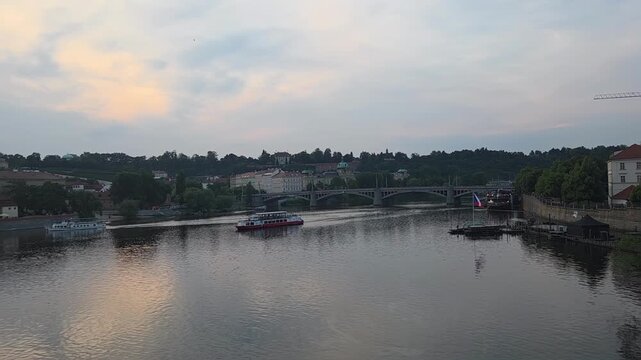Panning tracking shot of river boat cruising along historic riverside past bridges and buildings at dusk. Prague view of the Vltava