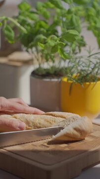 Hand slices bread with knife on cutting board, with potted herbs in the background. Concept of home cooking and recipe preparation