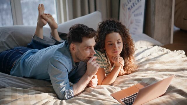 Couple browsing laptop lying cozy bed. Young pair relaxing weekend home morning.