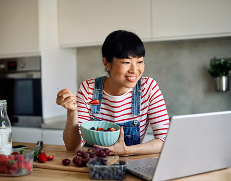 Portrait of a healthy young asian woman enjoying delicious fruit and cereal salad breakfast bowl and using a laptop to work at home