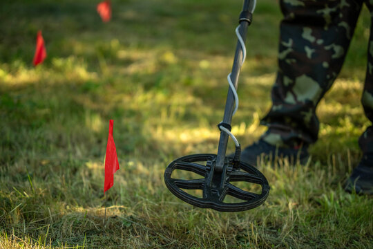A treasure hunter uses a metal detector to search for buried items in a grassy field. Small red flags mark spots of interest in the ground. The activity takes place during the daytime