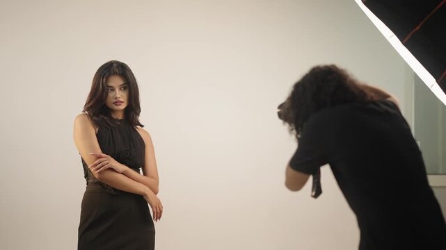 Close behind the scenes view of photographer adjusting camera while brunette fashion model poses confidently in black dress under studio softbox light