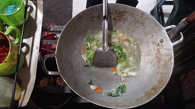High angle shot of a street food chef adding sauce and stirring fresh mixed vegetables in a large wok, cooking traditional Indonesian capcay.