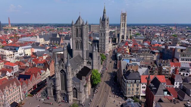 Aerial wide angle panoramic view of the three famous historic towers of Ghent aligned in a row along a city street with tram tracks under a bright summer sky