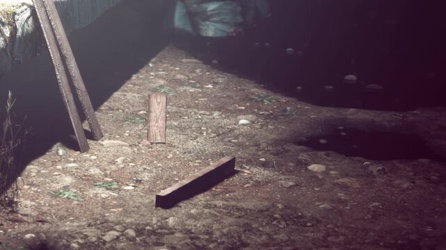 A person is hiding under a wooden structure. The ground has dirt and small plants. A few stones are scattered around. The atmosphere is unclear due to shadows and limited light.