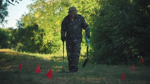 Male metal detectorist in camouflage attire searches for buried objects in a grassy area marked with red flags, surrounded by lush greenery