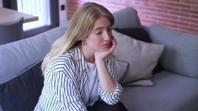 A young woman sits alone on a cozy sofa at home, looking sad and contemplative. Her pensive expression suggests feelings of boredom, loneliness, or emotional distress, appearing lost in thought or dea