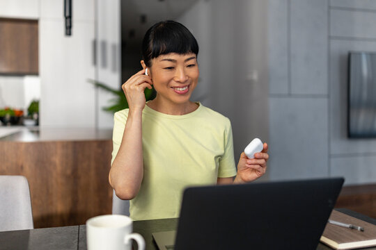 Japanese woman smiling during video call at home with earbuds and laptop in modern kitchen
