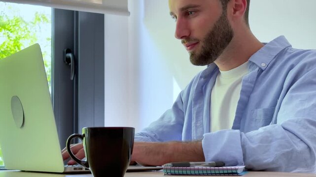 A focused young man working on his laptop, taking notes, and smiling at his home office desk. He enjoys a hot drink while engaged in online tasks, representing productivity, modern work, and a balance