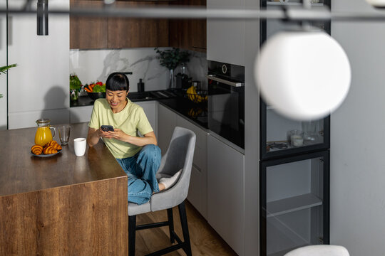 Japanese woman sitting at kitchen island smiling and using smartphone during relaxed breakfast