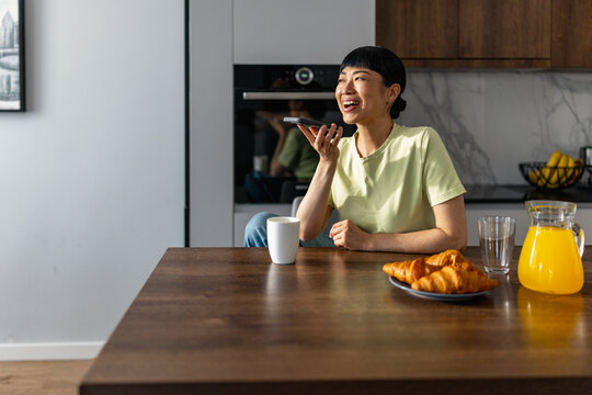 Japanese woman laughing while voice messaging at kitchen table with coffee and croissants
