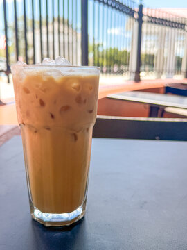 Glass of iced coffee with ice cubes sitting on an outdoor cafe table with blurred railing and seating in the background.