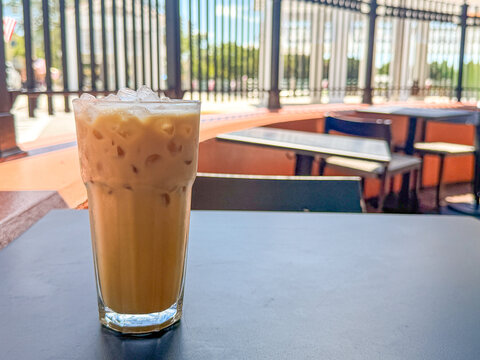 Glass of iced coffee with ice cubes on an outdoor patio table with empty cafe seating and railings in the background.