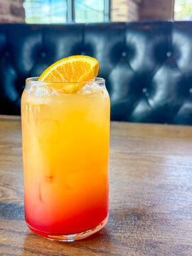 Colorful orange and red iced drink with citrus garnish served in a glass on a wooden table inside a restaurant booth.