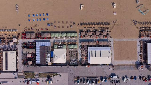 Aerial top down view of a row of summer beach clubs and restaurant pavilions next to a paved promenade and parking lot on the sandy coast of Scheveningen in The Hague