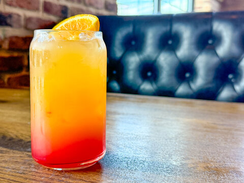 Bright orange and red iced beverage with orange slice garnish on a wooden restaurant table with booth seating in the background.