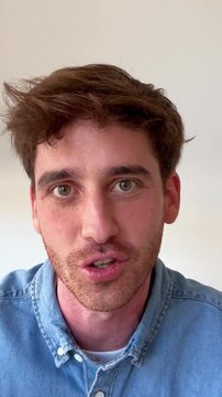A young man with brown hair and a beard, wearing a denim shirt, looks up with surprised and happy expressions, smiling and reacting to something off-camera with a sense of wonder and amazement.