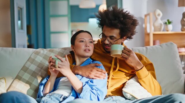 Multiracial couple relaxing on sofa drinking coffee watching TV together at home
