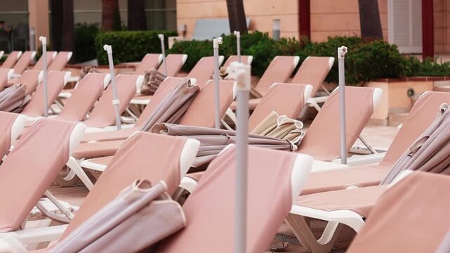 Rows of empty lounge chairs with beige cushions and white frames arranged neatly by a swimming pool area surrounded by greenery and modern architecture