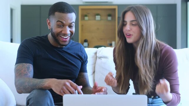 A happy mixed-race couple sitting on a cozy sofa at home, joyfully using a laptop together. They react with excitement and celebrate with a high-five, sharing a moment of success and good news.