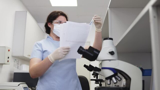 Medical laboratory technician wearing a mask and gloves checks a blood sample tube and compares the information with laboratory documents beside a microscope.