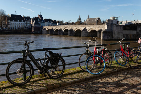 Maastricht, Netherlands - February 25, 2026: Bicycles parked along the Maas River embankment with the historic Sint Servaasbrug stone bridge and cityscape in the background under a clear blue sky
