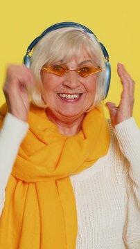 Excited senior stylish old woman listening music via headphones, dancing disco party fooling around having fun expressive gesticulating hands. Happy elderly grandmother pensioner on yellow background