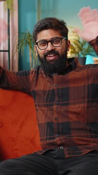 Hello. Indian handsome man smiling friendly at camera and waving hands gesturing hello hi, greeting or goodbye welcoming with hospitable expression at home. Arabian guy sitting on sofa in living room
