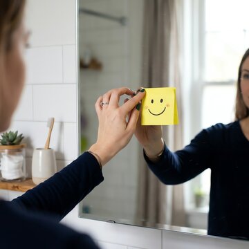 Woman sticking a smiling face sticky note on bathroom mirror for positive affirmation