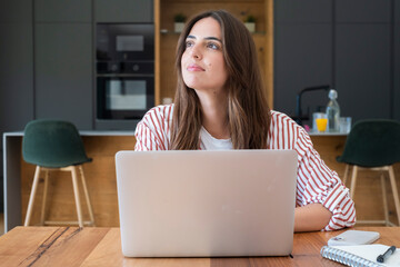 Young woman working on a laptop in a modern kitchen, looking thoughtful and planning ideas while doing remote work with a notebook, phone and glass of orange juice nearby © simona
