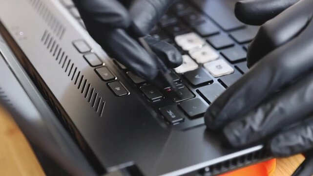 Hands of one young Caucasian guy in black latex gloves glues transparent stickers with a letter and a number on the laptop keyboard buttons with special tweezers, sitting at the office desk in the bed