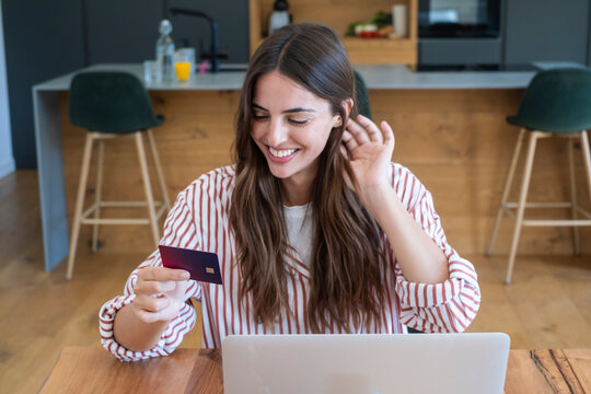Young woman smiling while shopping online with a credit card at her laptop in a modern kitchen, making an ecommerce payment and enjoying convenient home shopping