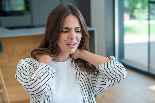 Young woman holding her neck in pain and discomfort at home, grimacing from neck ache and tension while seeking relief and self-care in a bright modern living space