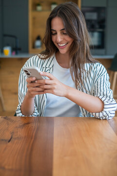 Young woman smiling and using smartphone at wooden table in modern kitchen, casually dressed, texting or browsing, relaxed mood and natural lifestyle scene
