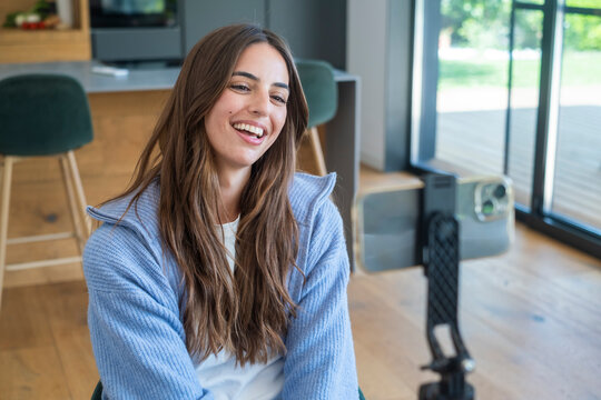 Smiling young woman recording a vlog on smartphone at home, casually dressed in a blue sweater, engaging with audience while sitting in a bright kitchen-living area, lifestyle content creator 