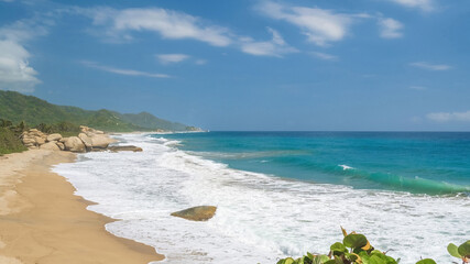 Obraz premium Horizontal image. Tropical beach with waves breaking on sand and large smooth rocks under a blue sky
