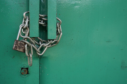 A close-up shot of an old rusty padlock and a heavy steel chain wrapped tightly to lock a green painted metal door.                               