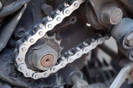 A detailed close-up shot of a metallic motorcycle drive chain installed around a dirty and greasy front engine sprocket with a rusty nut.                               