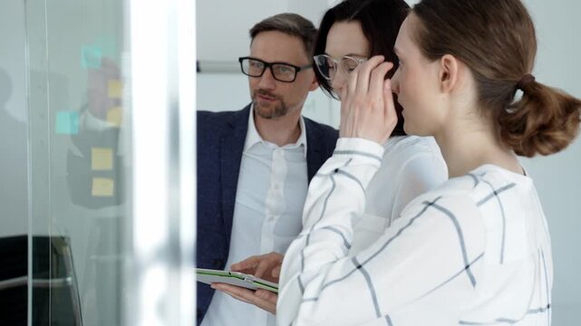 Business team collaborating on a project, analyzing data on a tablet, planning strategy with sticky notes on a glass whiteboard in a modern office meeting