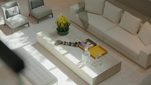 Bright modern living room with light sofa and matching armchairs, large wooden coffee table in center holds yellow flowers, books, and decor