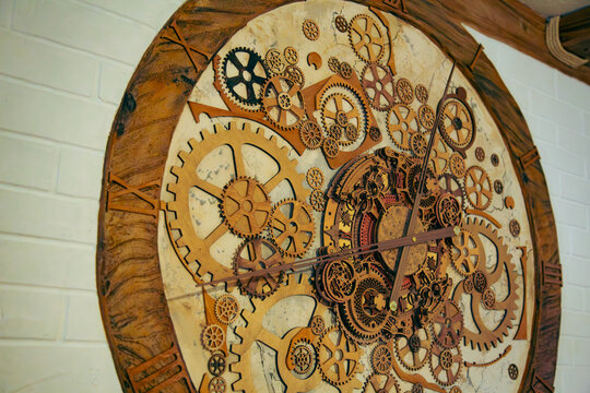 This beautiful interior photograph captures a large decorative steampunk wall clock made of natural wood shot from a creative three quarter angle. The intricate mechanical design features numerous