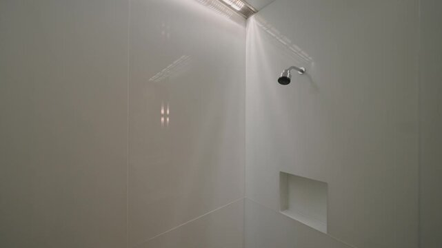 Clean shower space with smooth light walls, small shelf built in wall, silver showerhead above, glass door reflects soft light, room looks fresh and simple