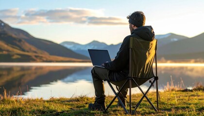Fototapeta premium Man working on laptop while sitting in folding chair by mountain lake at sunrise, remote work lifestyle