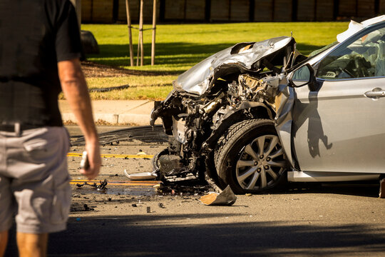The front end of a silver car heavily damaged at the scene of an auto accident. A man is in the foreground