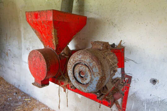 Rusty red agricultural machine mounted on white barn wall with dirt and straw floor